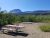 Red picnic table and small grill on dry ground with green shrubs, distant mountains in Glacier National Park.