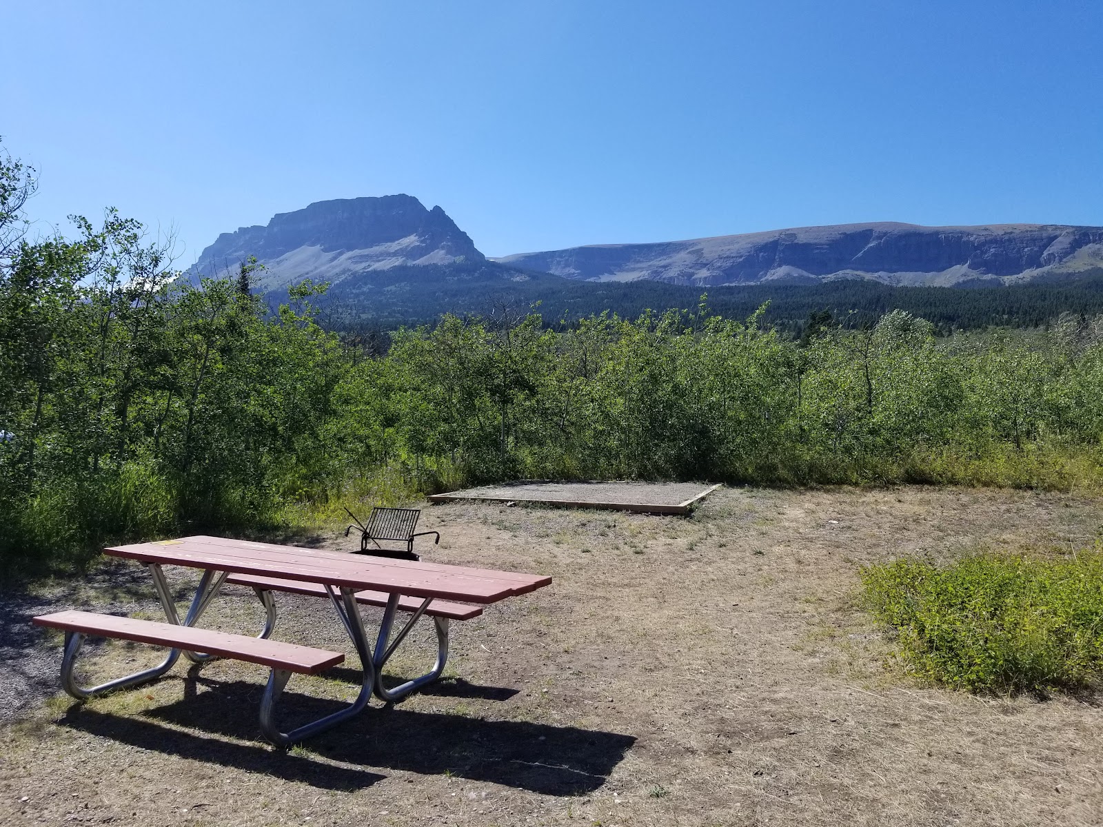Red picnic table and small grill on dry ground with green shrubs, distant mountains in Glacier National Park.