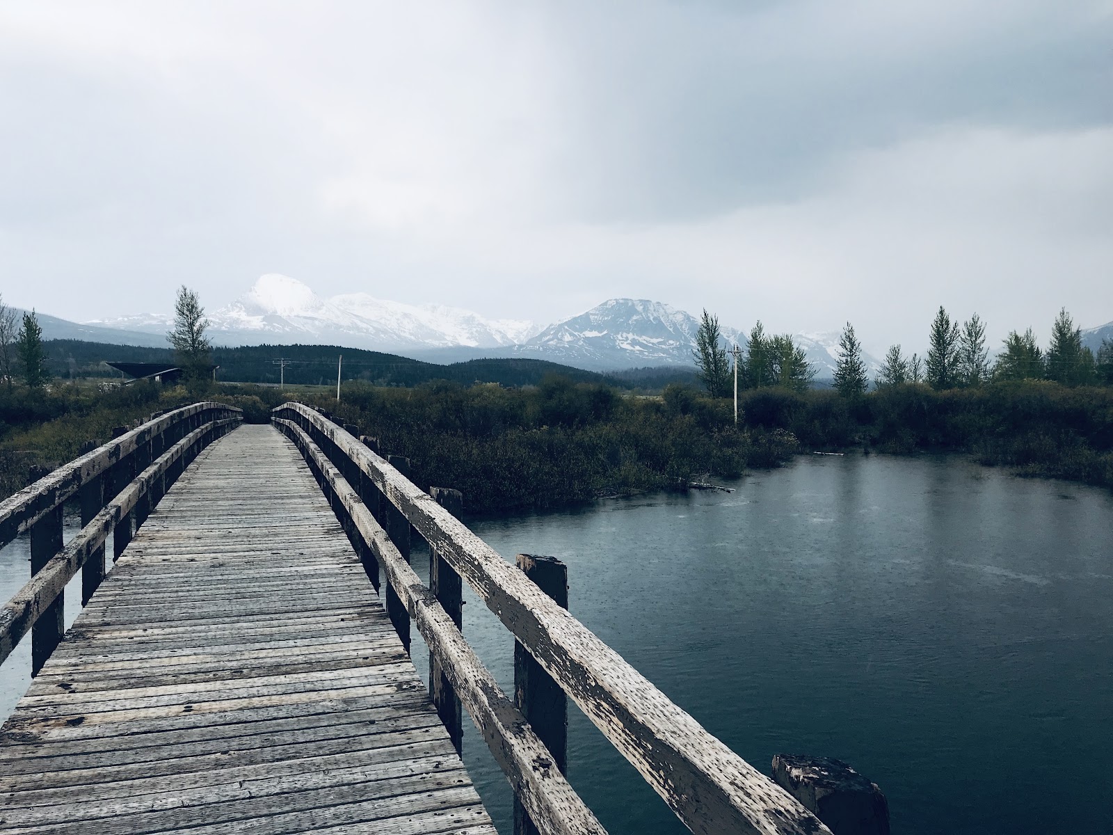 Weathered wooden boardwalk over a calm lake leads toward snow‑capped mountains and pines in Glacier National Park.