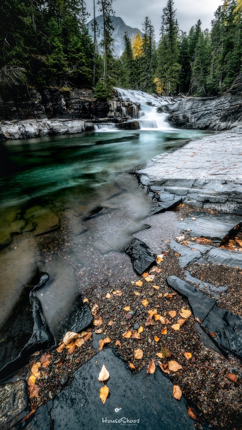 John's Lake waterfall scene in Glacier National Park with emerald pool, rocky shoreline, and autumn foliage along the forested valley.