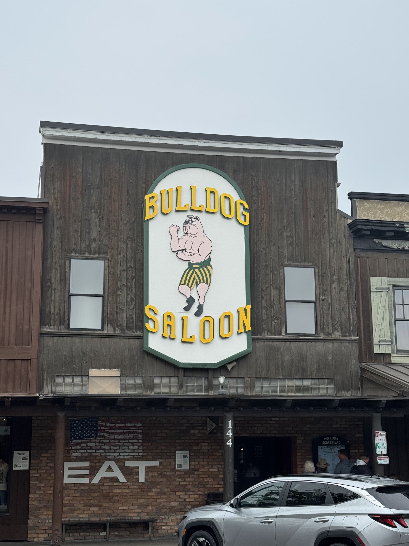 Rustic wooden storefront with a bold sign for a saloon in Glacier National Park, seen with a brick base and people nearby.
