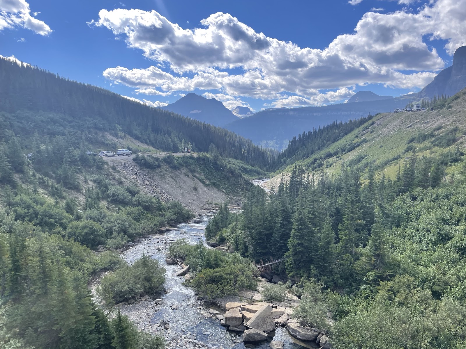 Glacier National Park valley along Siyeh Pass Trail featuring a rocky stream, dense evergreens, and distant rugged peaks under a bright sky.