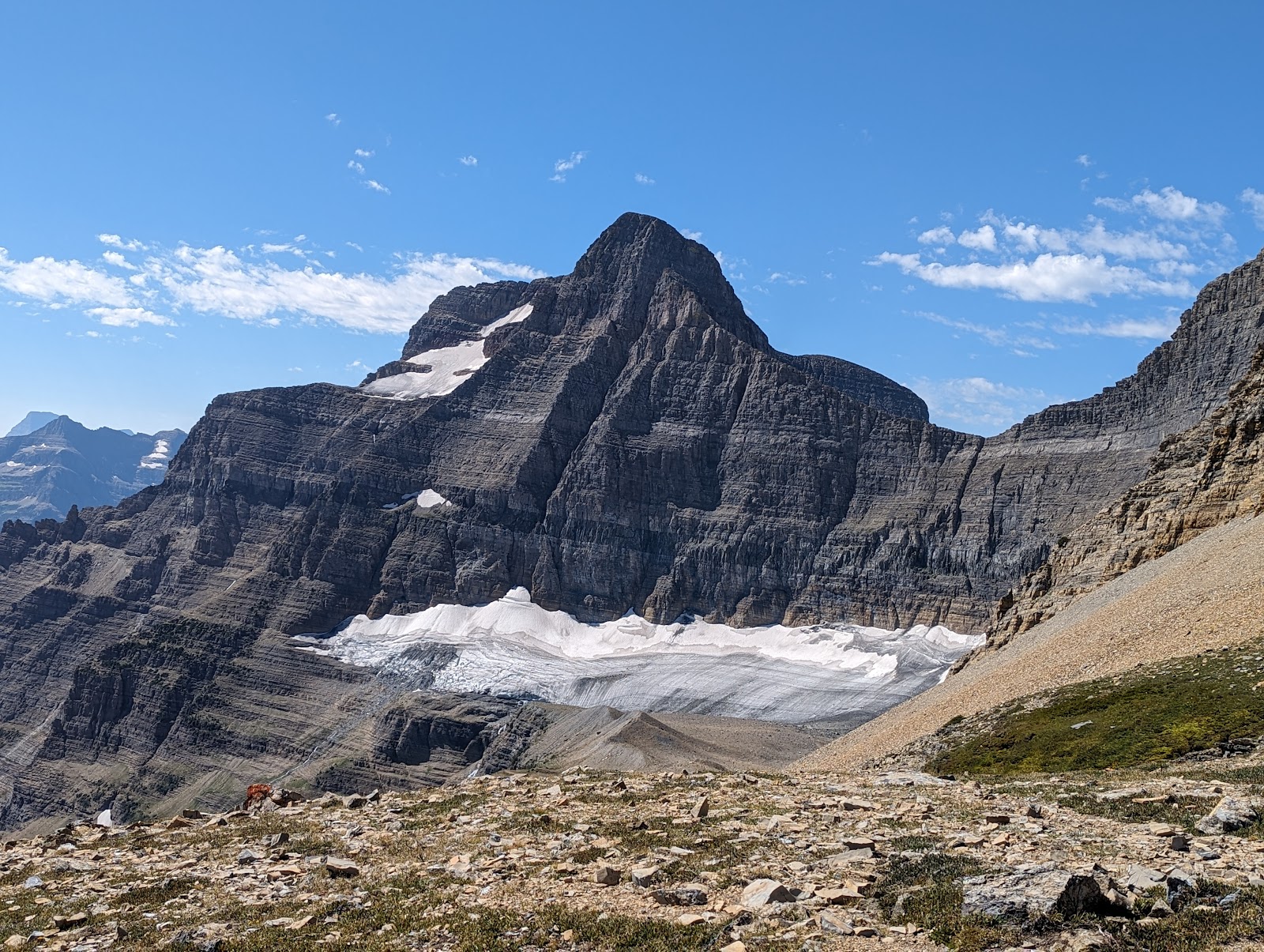 Jagged granite peak with snow patches and a broad glacier in a rocky valley under a blue sky, Glacier National Park.
