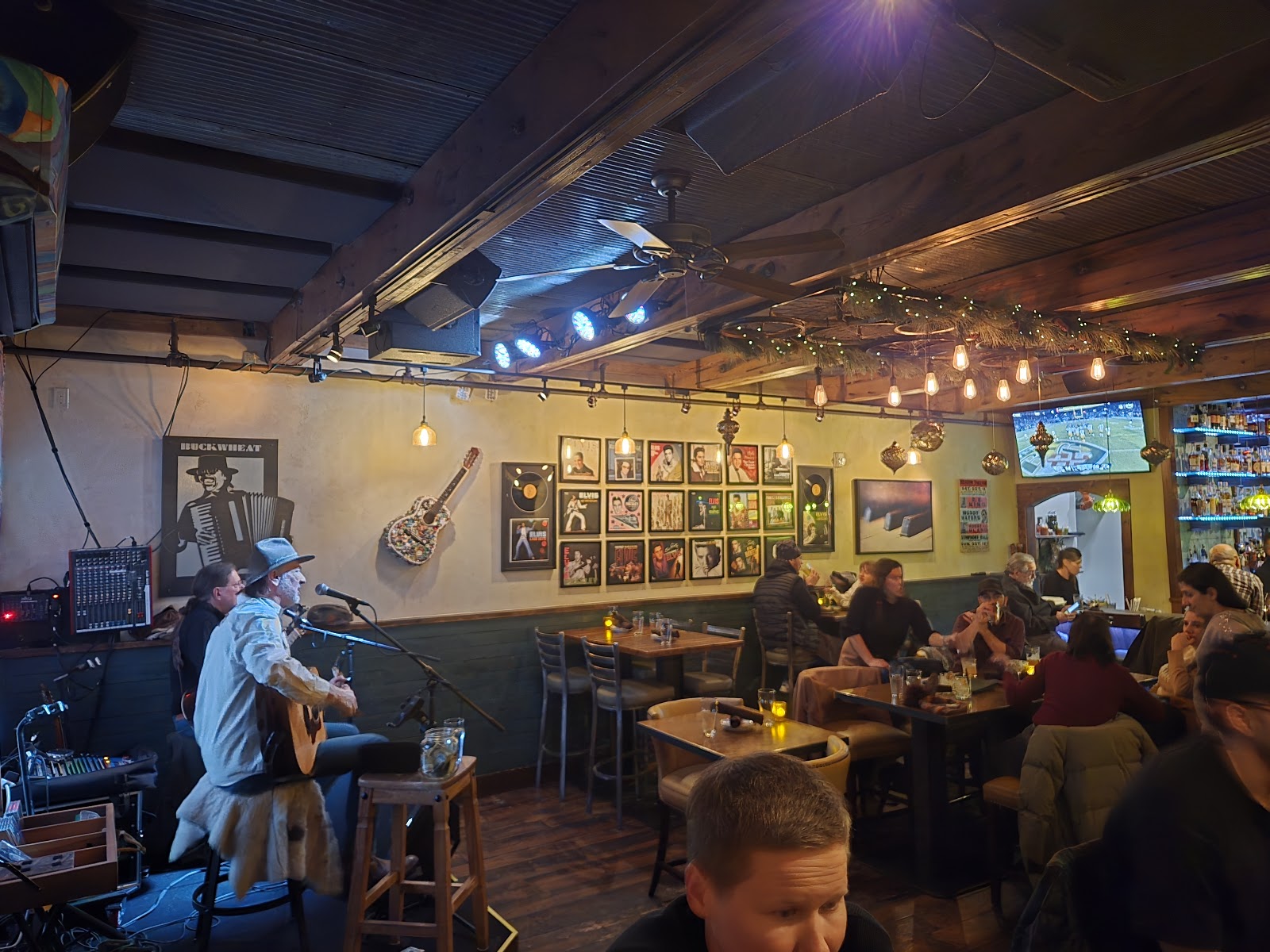 Cozy Glacier National Park restaurant interior with a guitarist on stage, diners at tables, warm hanging lights, and wall art.