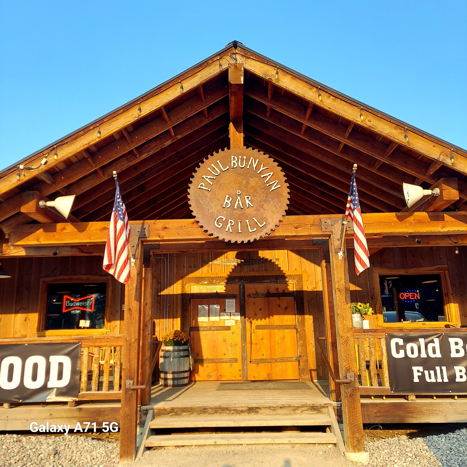 Rustic lodge-style bar and grill exterior at Glacier National Park with a round sign and American flags flanking the entry.