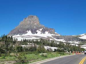 Rugged mountain with patches of snow rises over evergreen trees; a road, orange cones, and a van in Glacier National Park.