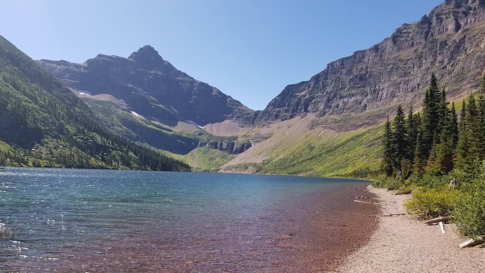 Clear blue Two Medicine Lake reflects rugged glacier peaks and evergreen slopes along a pebbled shore in Glacier National Park.