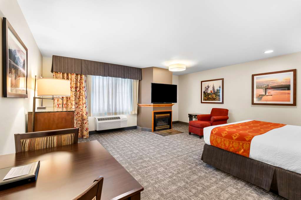 Spacious Glacier National Park hotel room with a bed and orange throw, red armchair, fireplace, wall art, TV, desk and patterned curtains.
