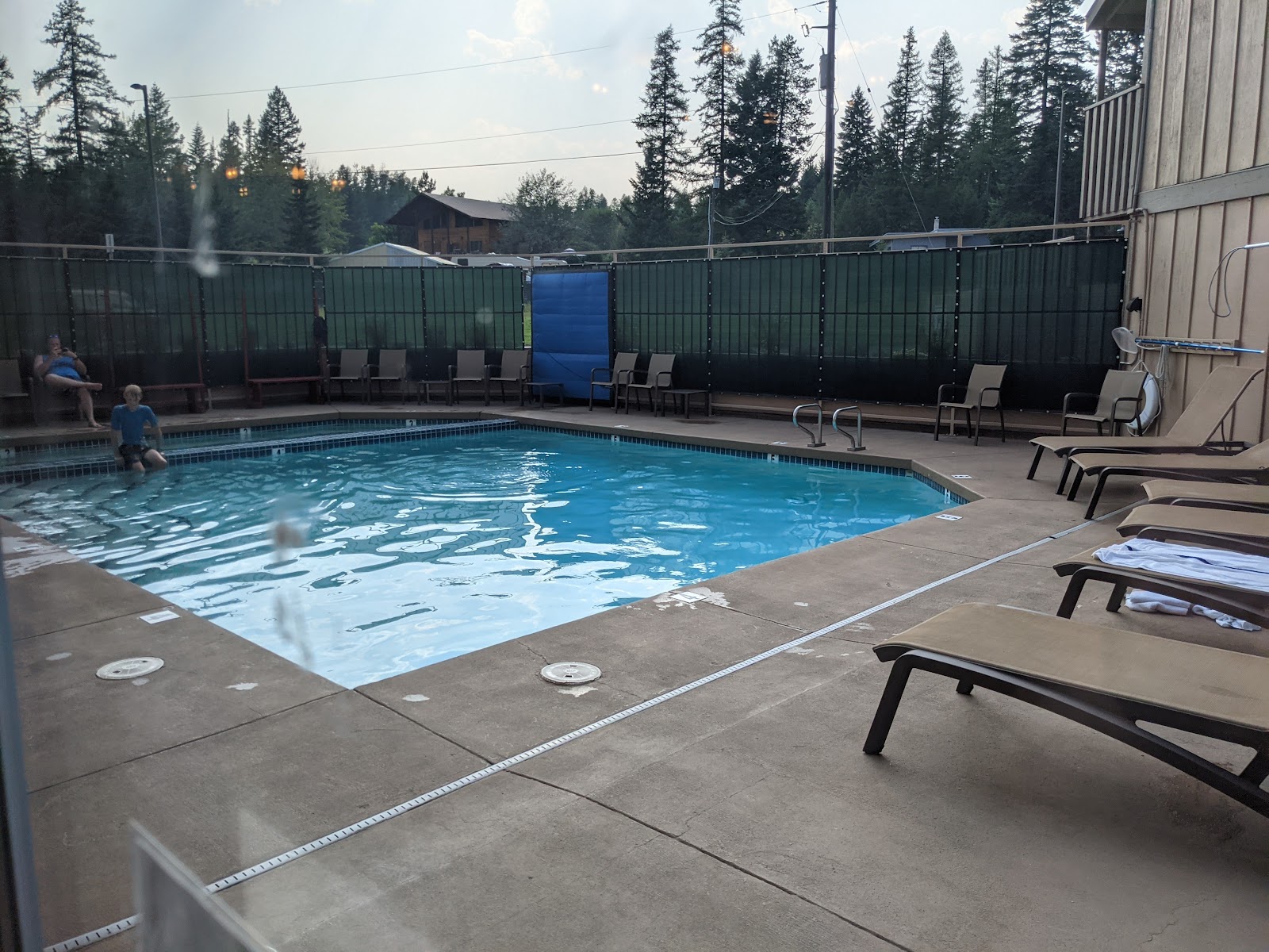 Lodge pool area with blue water, tan lounge chairs, and a green privacy fence against evergreen trees in Glacier National Park.