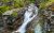 Rugged granite cliffs frame a rushing waterfall at Siyeh Pass Trailhead in Glacier National Park, surrounded by moss and pines.
