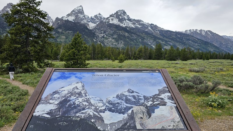 Signboard at Teton Glacier Turnout in Yellowstone National Park with snow-capped peaks, pines, and a hiker.