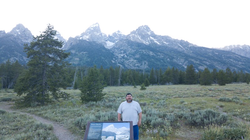 Man stands beside an informational sign at a turnout in Yellowstone National Park, with sagebrush and snow‑capped mountains in the background.