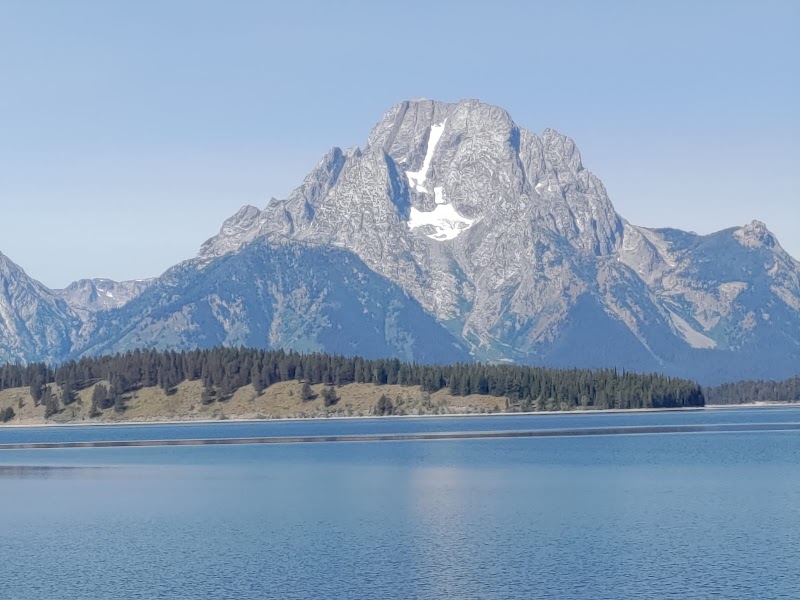 Snow-dusted jagged peak rises above a forested shoreline and calm blue lake in Yellowstone National Park.