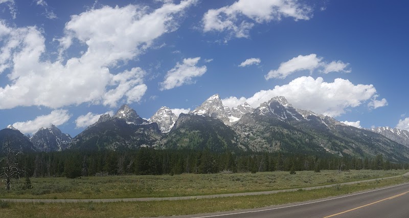 Snow-capped peaks of the Teton Range rise behind a pine forest, with a two-lane road in the foreground at Yellowstone National Park.