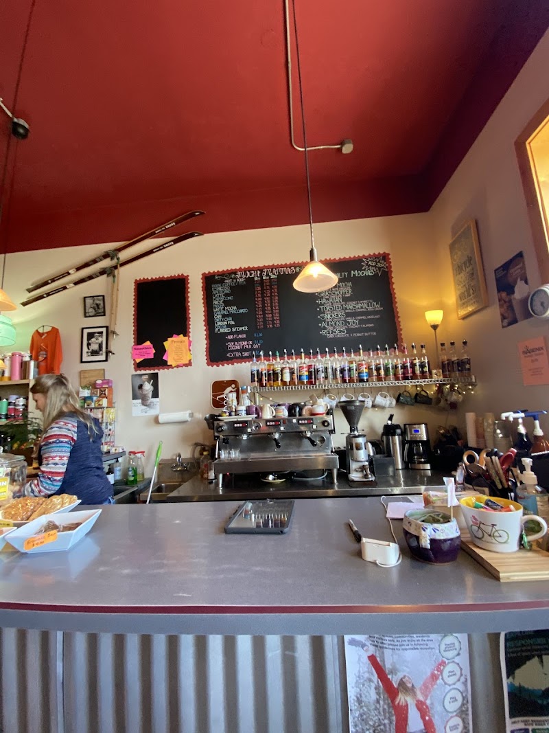 Inside a cozy cafe in Yellowstone National Park, stainless counter, espresso machine, chalkboard menu, hanging skis, and a staff member at left.