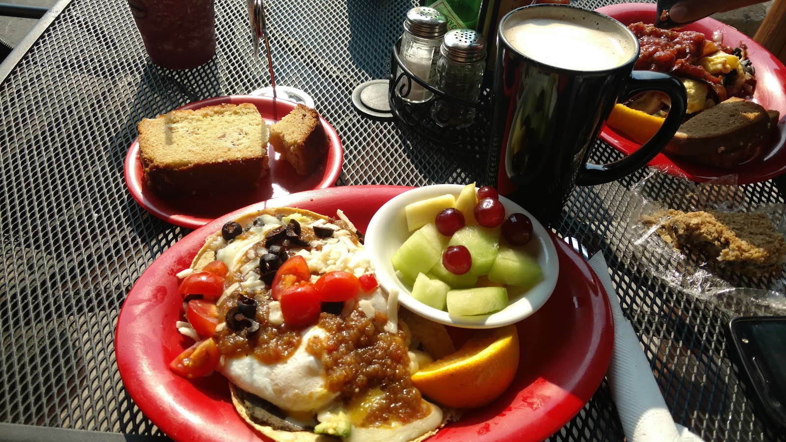Glacier National Park picnic: red plate with a loaded taco, a bowl of melon and grapes, orange slice, bread slices, and a tall black coffee mug on a mesh table.