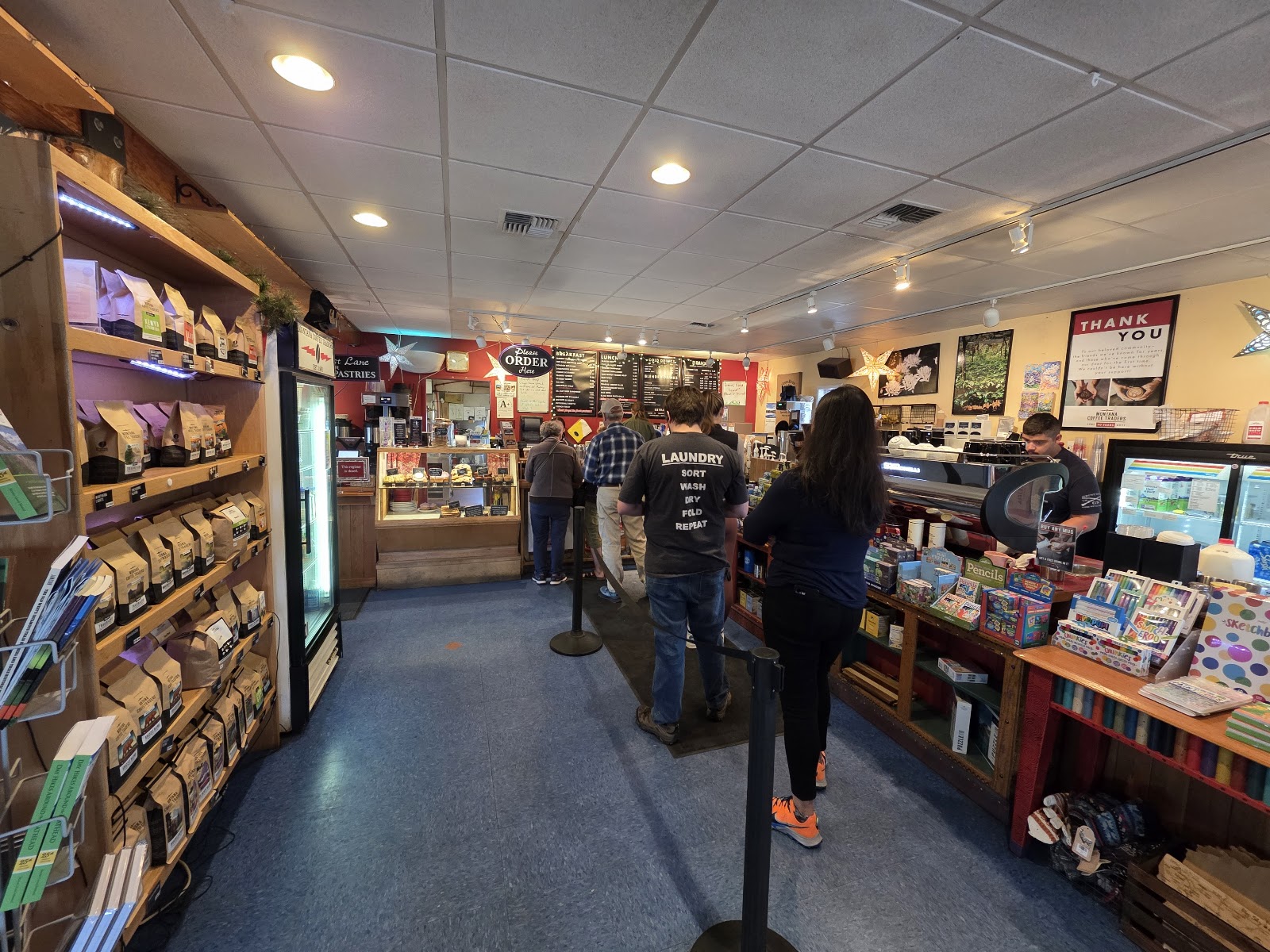 Cozy cafe interior in Glacier National Park with wooden shelves of coffee bags on the left, a glass display case and menu boards at the back, and customers waiting in line.