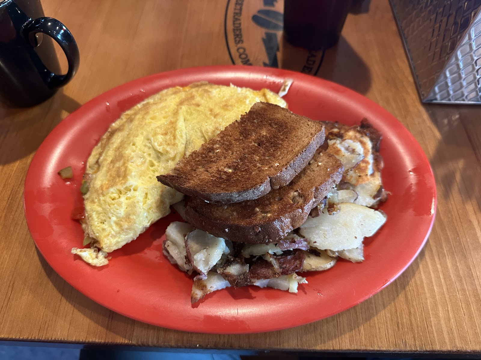 On a wooden table, a red plate holds a folded omelette, dark rye toast, and hash browns with bacon in Glacier National Park.