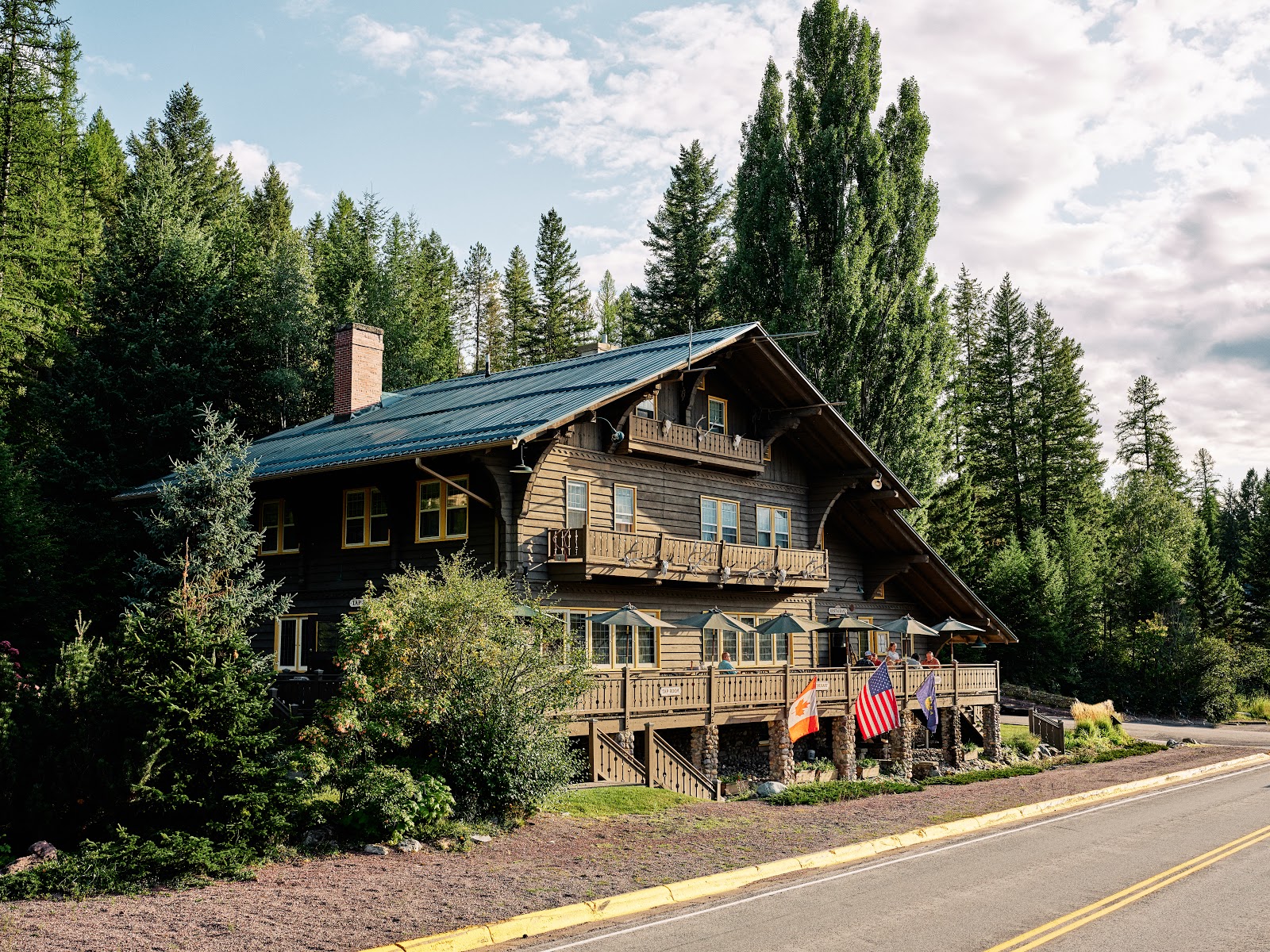 Chalet-style lodge along a forested Glacier National Park road, with a wooden deck, flags, and dense pines nearby.