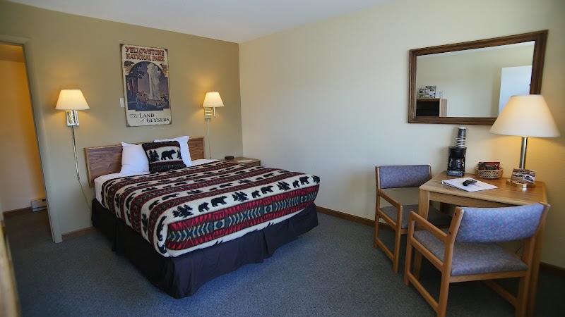 Cozy Yellowstone National Park motel room with a king bed, patterned quilt, two wall lamps, a wooden desk and chairs, and a wall mirror.
