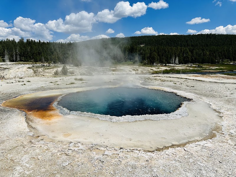 Castle Geyser hot spring in Yellowstone National Park, a blue crater pool with steam, orange mineral rims, and forested hills beyond.