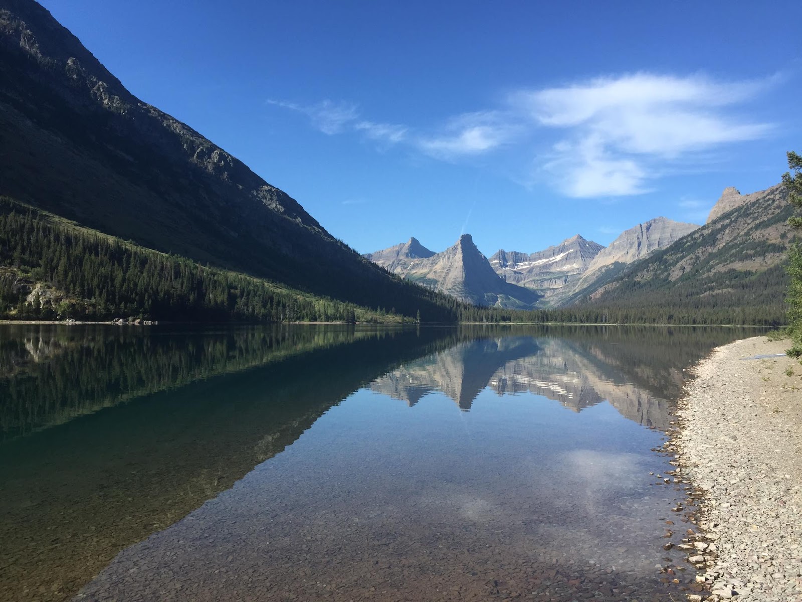 Calm lake reflecting Pyramid Peak and jagged glacier mountains, with a pine forest along the shore in Glacier National Park.