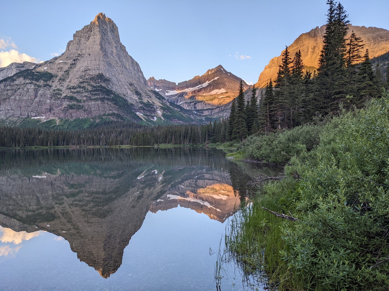 Pyramid Peak rises over a glassy lake in Glacier National Park, with snow patches, evergreen forest, and a perfect reflection.