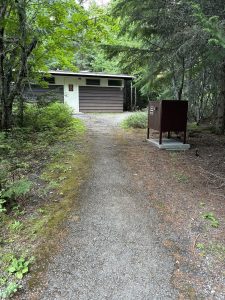 Gravel path leads to a small brown building with white door amid green trees, plus a brown storage box on a concrete pad in Glacier National Park.