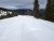 Snowy slope with several skiers beside a sign, evergreen trees, and distant snow‑capped peaks in Glacier National Park.