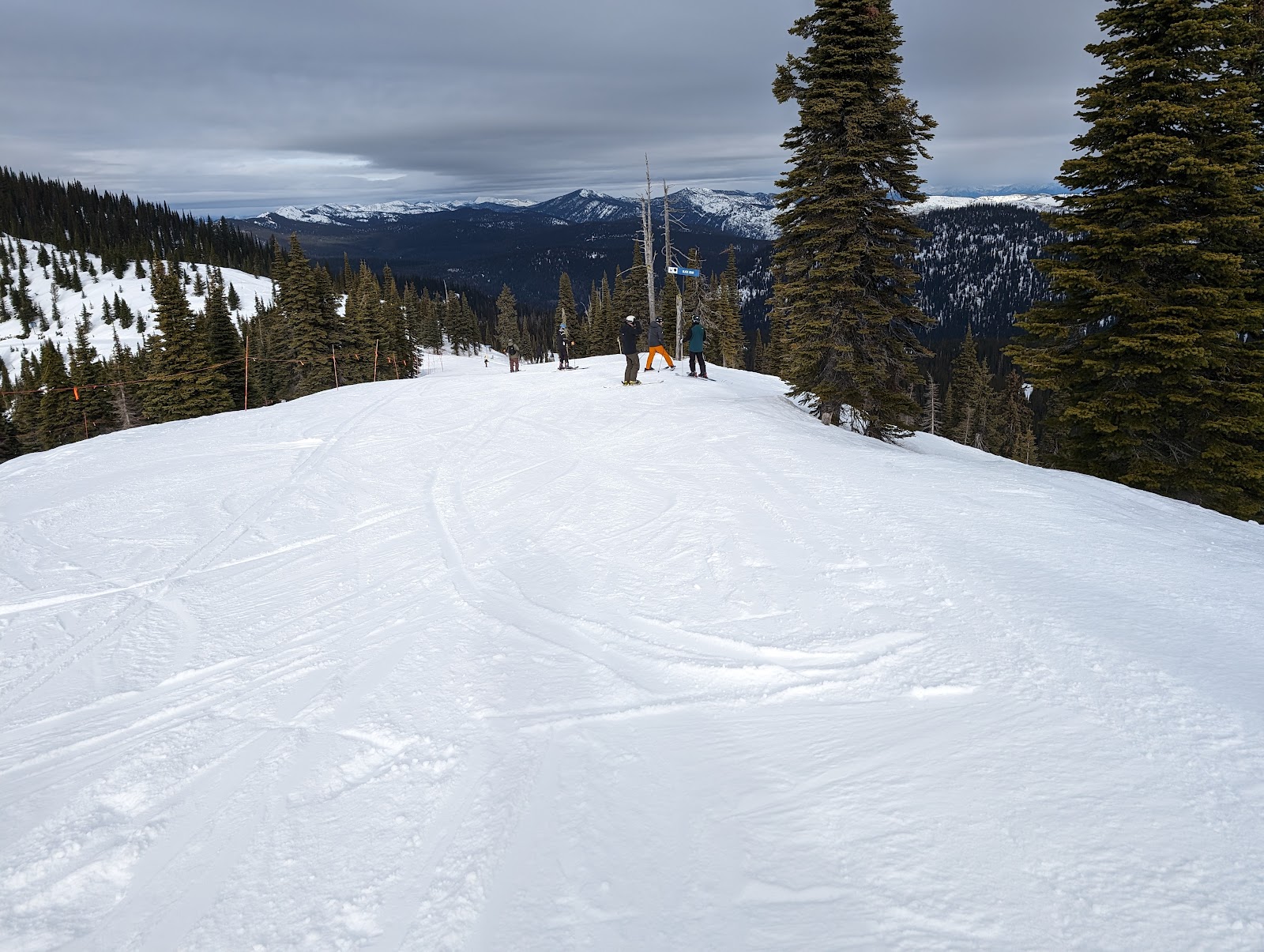 Snowy slope with several skiers beside a sign, evergreen trees, and distant snow‑capped peaks in Glacier National Park.