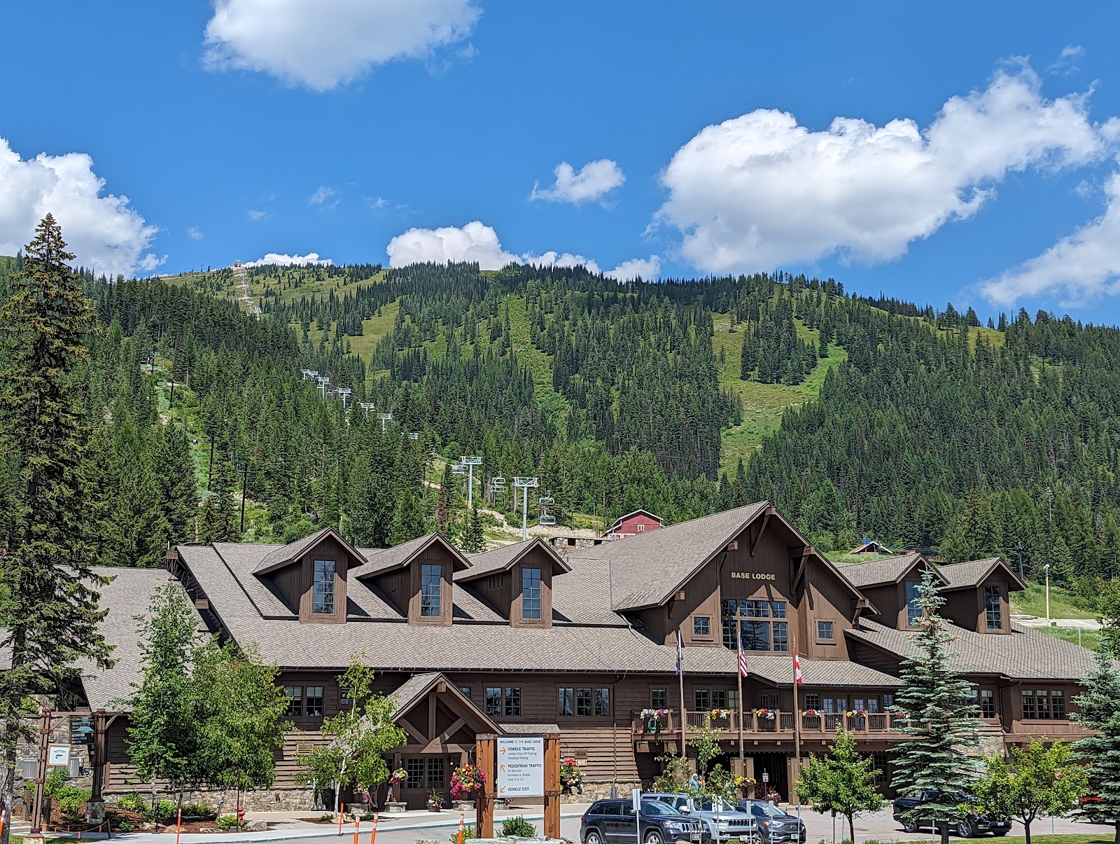 Wooden lodge with gabled roofs and balconies sits at Glacier National Park's evergreen hillside under a bright blue sky.