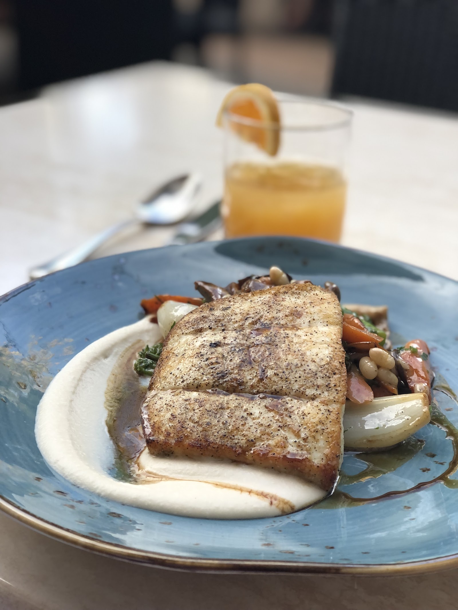 Seared rectangular fish fillet on a blue plate with creamy white sauce and sautéed vegetables, plus an orange cocktail with a lemon garnish in Glacier National Park.