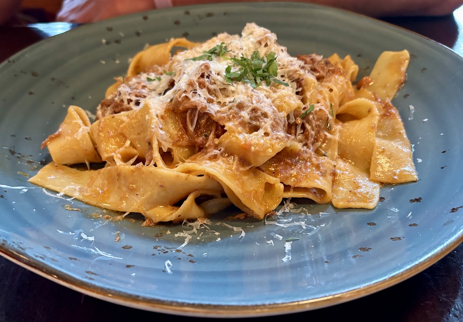 Plate of wide pappardelle pasta with meat sauce, grated cheese, and parsley on a blue ceramic plate in Glacier National Park.