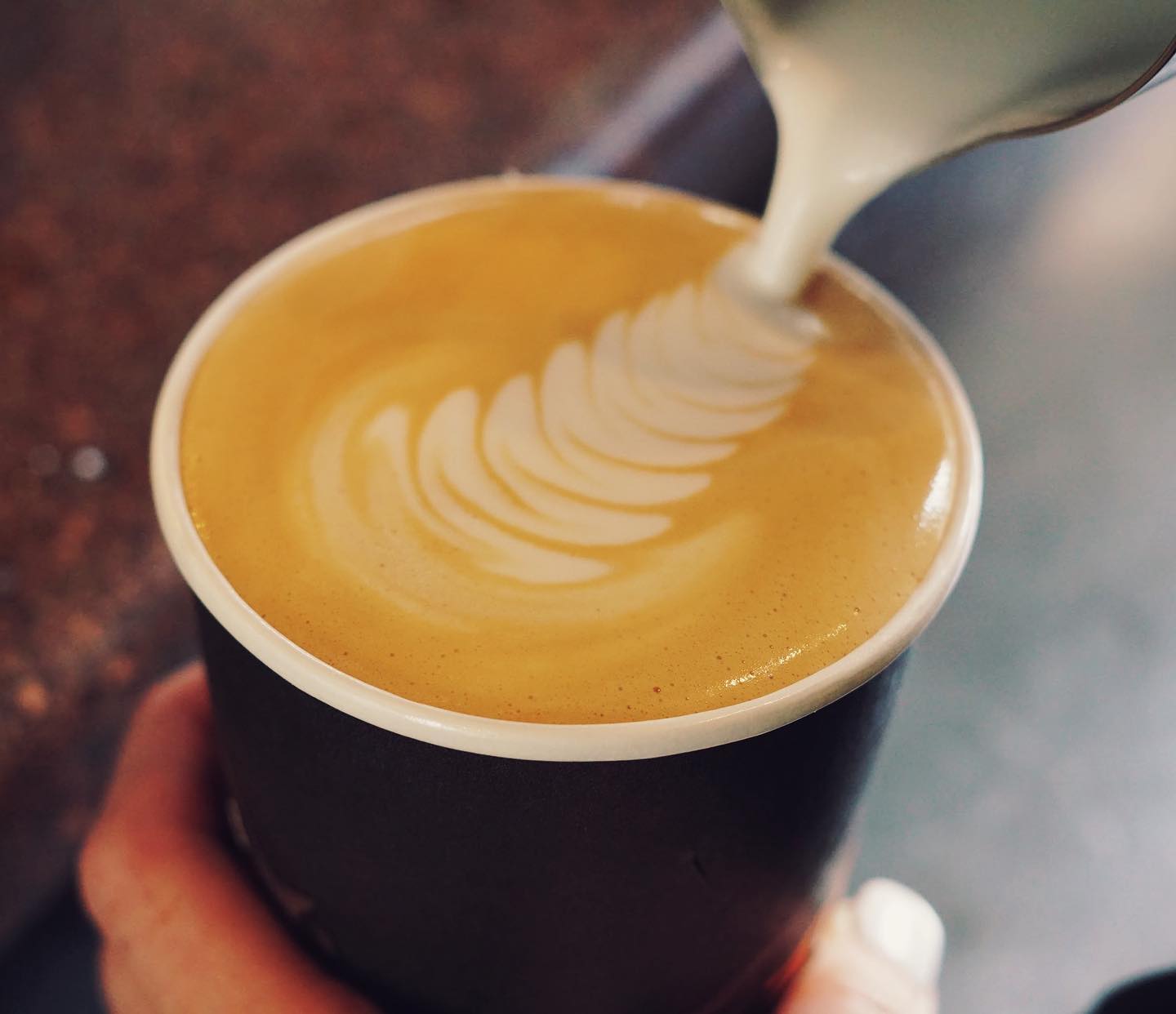 Latte with intricate latte art being poured into a dark disposable cup at a Glacier National Park cafe.