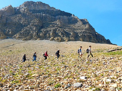 Matahpi Peak trail hikers ascend a rocky slope beneath a towering granite face in Glacier National Park.