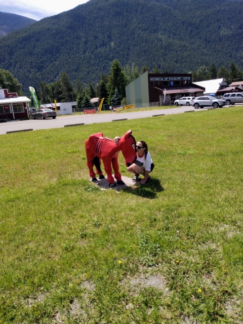 In Glacier National Park, a person kneels beside two red horse statues on a grassy area with buildings and forested mountains behind.