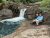 Deadwood Falls at Glacier National Park cascades between reddish rock walls as two hikers rest on a rocky ledge beside the pool.