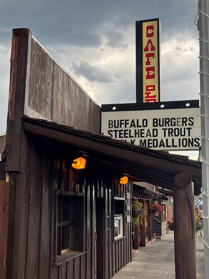 Rustic wooden storefront along a sidewalk in Yellowstone National Park, featuring a tall vertical cafe sign and a marquee menu.