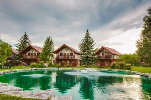 Lodge-style cabins line a turquoise pond at Glacier National Park, with evergreens and well-kept lawns in front of red-roofed chalets.