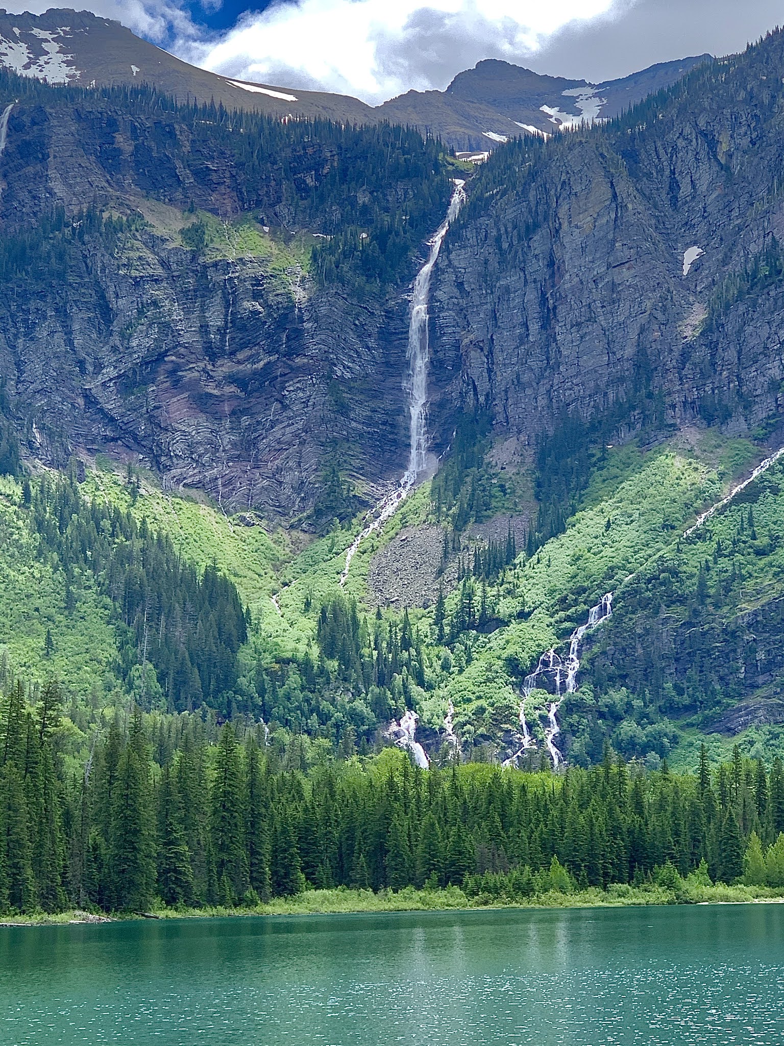 Gunsight Mountain waterfall cascades down rugged cliffs toward a turquoise lake in Glacier National Park.