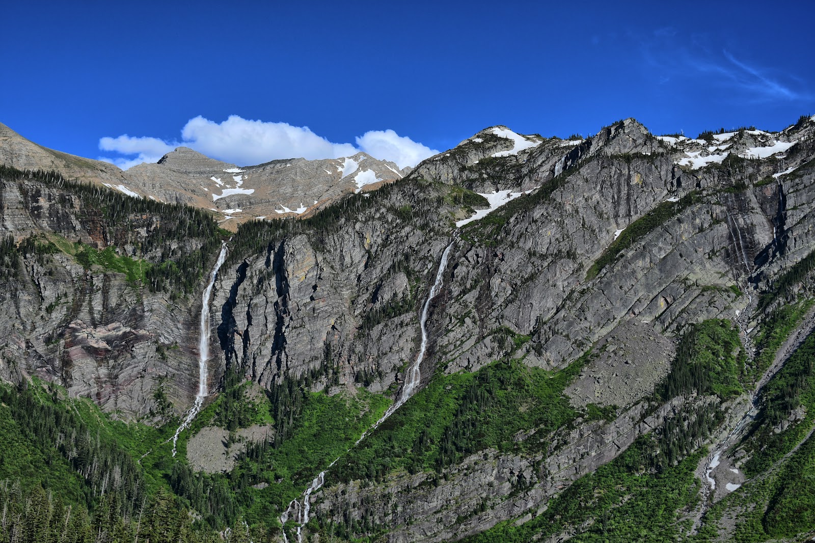 Gunsight Mountain rises above a rugged Glacier National Park valley with waterfalls and patches of lingering snow under a clear blue sky.