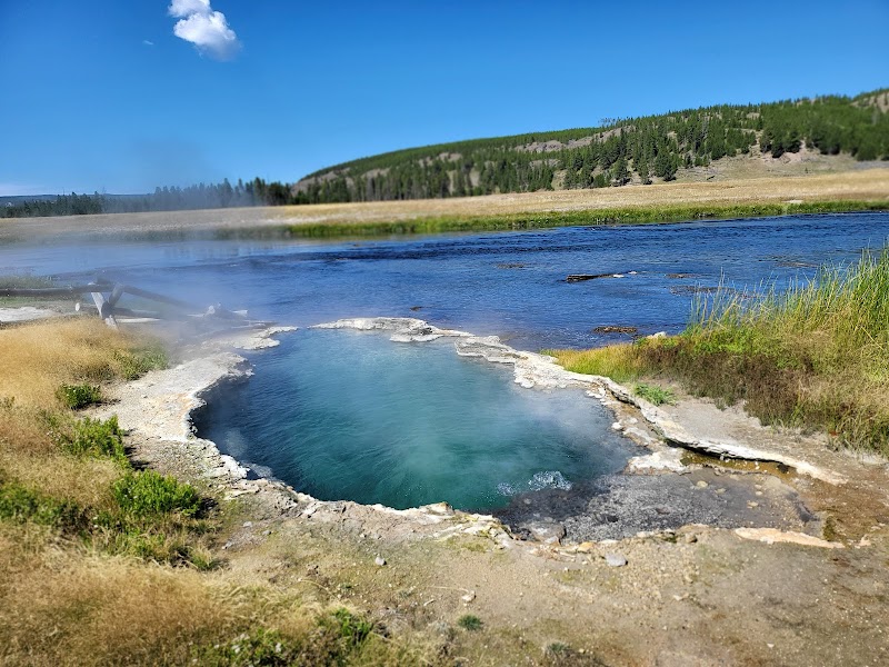 Steam from a blue mud pool with a mineral rim, grassy edges, and a distant river and hills in Yellowstone National Park.