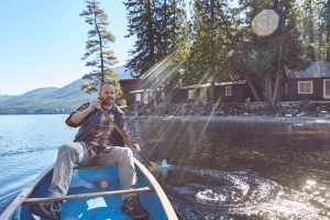 Canoe paddler in a blue canoe near Apgar Village on Lake McDonald, Glacier National Park, with forested shoreline and cabins.