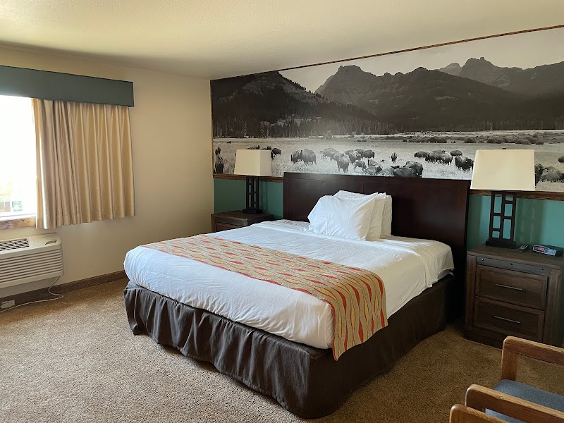 Yellowstone National Park hotel room with a king bed, white linens, orange runner, two nightstands with lamps, a window with curtains, and a mountain-bison mural