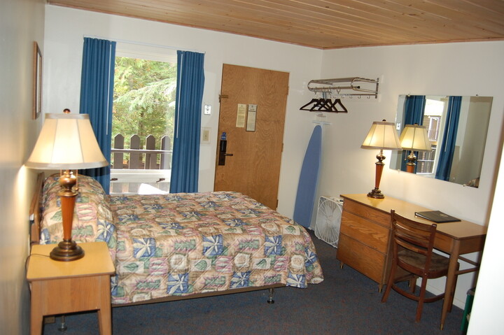 Cozy Glacier National Park motel room with a quilted bed, two lamps, wooden desk and dresser, blue curtains, door, and an outside view.