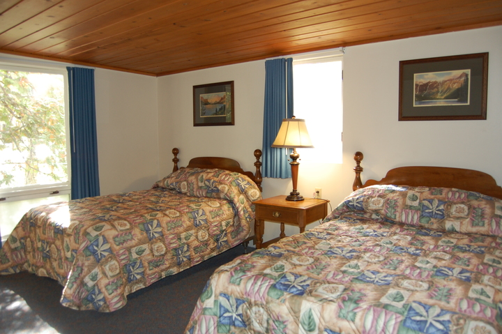 Two twin beds with colorful quilted bedspreads, a wooden nightstand and lamp between them, blue curtains and framed landscape photos in a Glacier National Park lodge room.