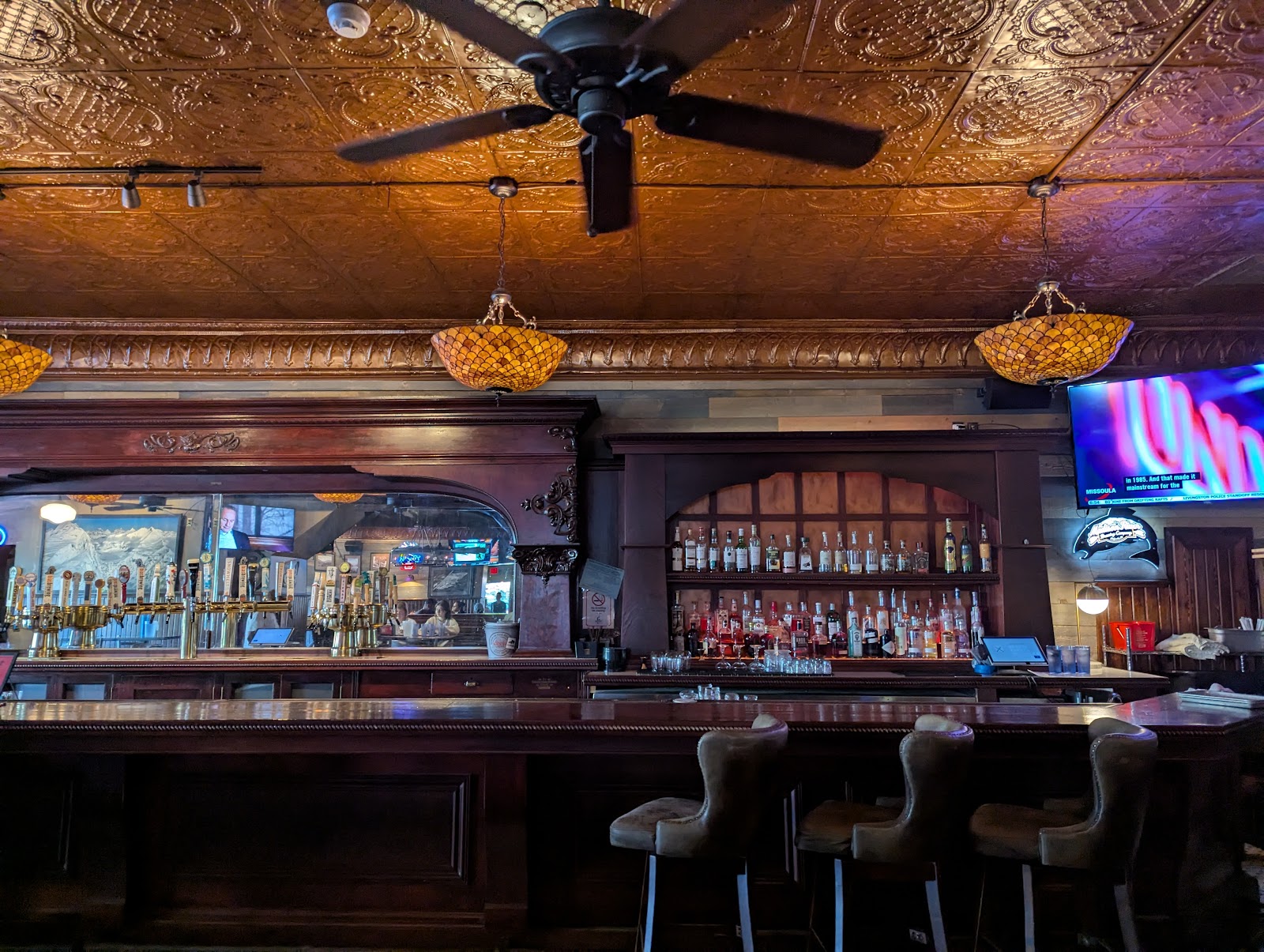 Interior of a historic saloon in Glacier National Park, featuring a dark wood bar, brass taps, and ornate copper ceiling.