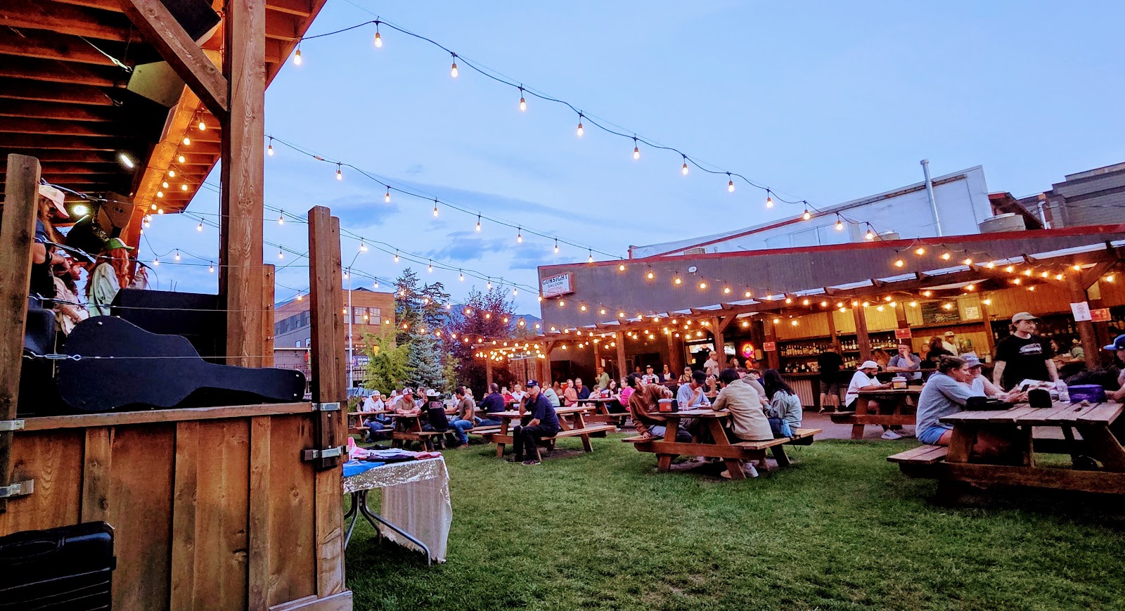 Rustic saloon courtyard at Glacier National Park lit by string lights, with numerous picnic tables and a lively evening crowd.