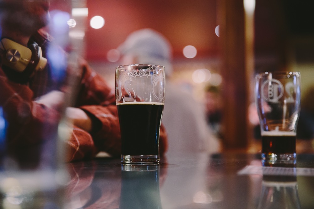 Two pints sit on a glossy bar at Bonsai Brewing Project near Glacier National Park, with warm lights and blurred patrons in the background.