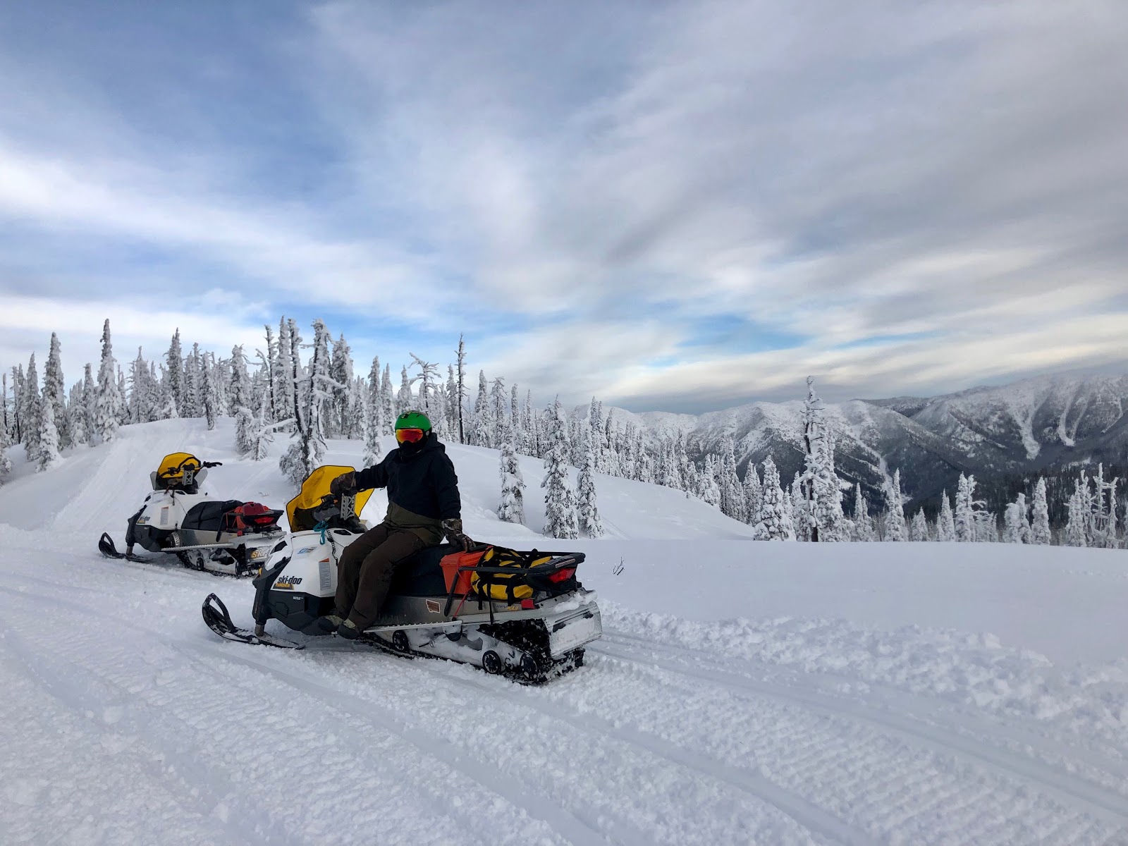 Snowmobiles on a snow-covered alpine ridge in Glacier National Park, with frost-coated trees and distant mountain peaks.